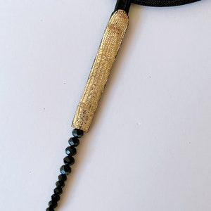 May include: A gold and black necklace with a long, thin pendant made of gold fabric and black beads.