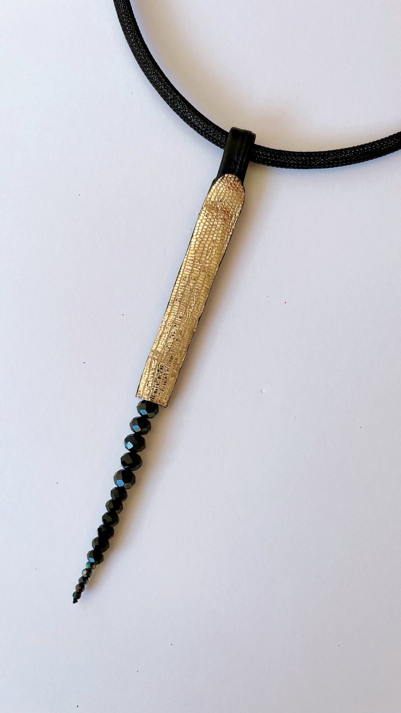May include: A gold and black necklace with a long, thin pendant made of gold fabric and black beads.