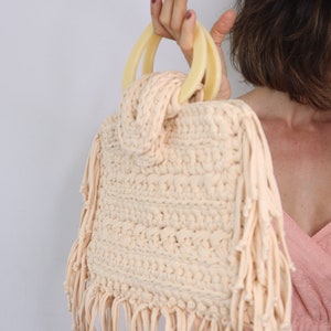 May include: A crocheted bag with fringe detailing and a light beige color. The bag has two large, cream-colored handles.