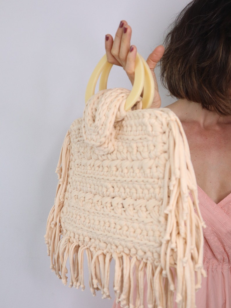 May include: A crocheted bag with fringe detailing and a light beige color. The bag has two large, cream-colored handles.