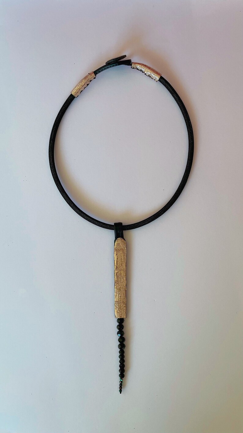 May include: A black choker necklace with gold accents and a pendant made of a gold-colored bar with a string of black beads hanging from it.