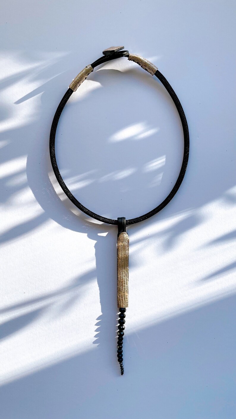 May include: A black choker necklace with a gold and black pendant. The pendant is a long, thin rectangle with a textured surface. The necklace is made of a thin, flexible material.