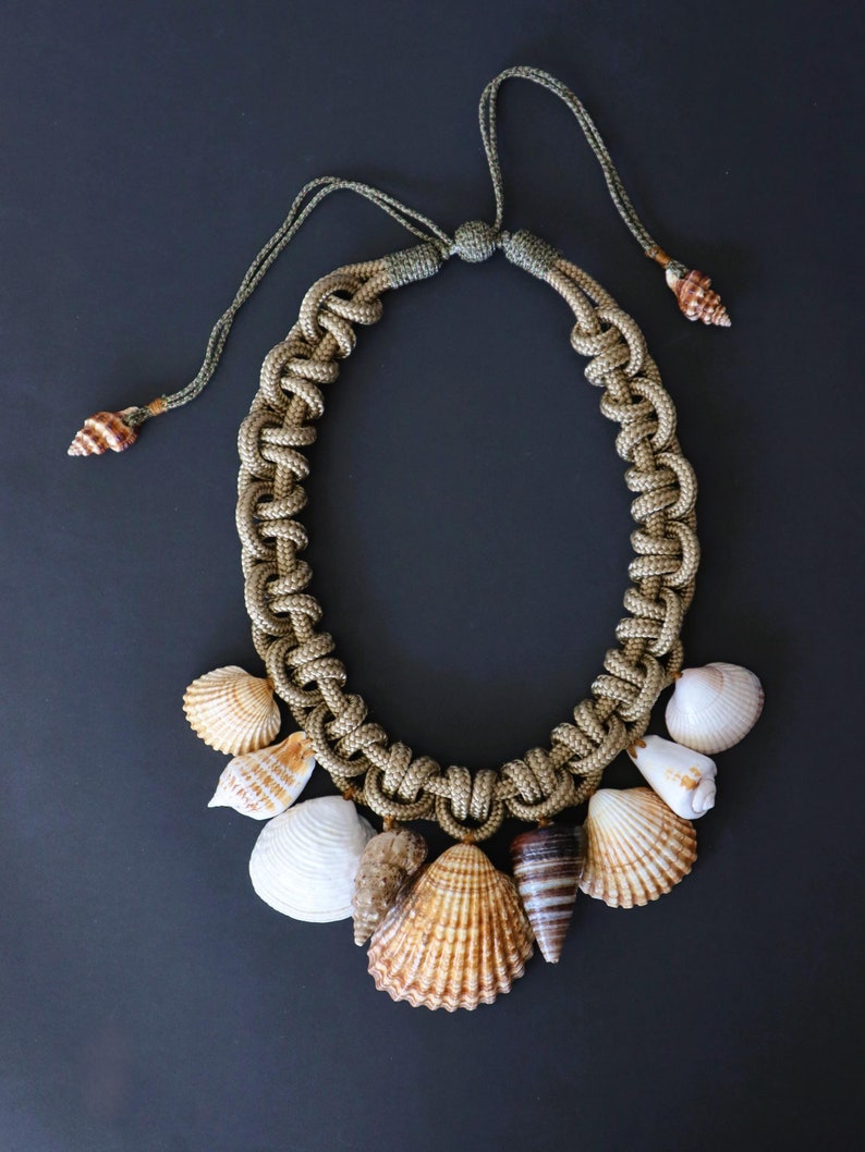 May include: A knotted rope necklace with a variety of seashells attached. The rope is a combination of green and brown colors. The seashells are a mix of white, brown, and tan.