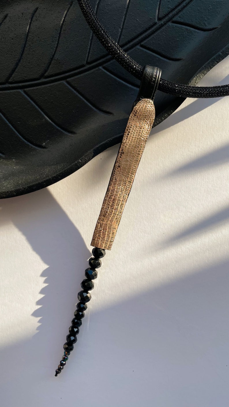 May include: A gold pendant necklace with a textured, rectangular charm and a long strand of black beads.