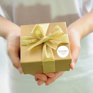 May include: A gold gift box with a gold ribbon and bow. The box has a white sticker with the text "VANESKA VILLA" on it.
