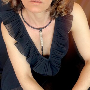 May include: A woman wearing a black ruffled dress with a long, silver and black pendant necklace.