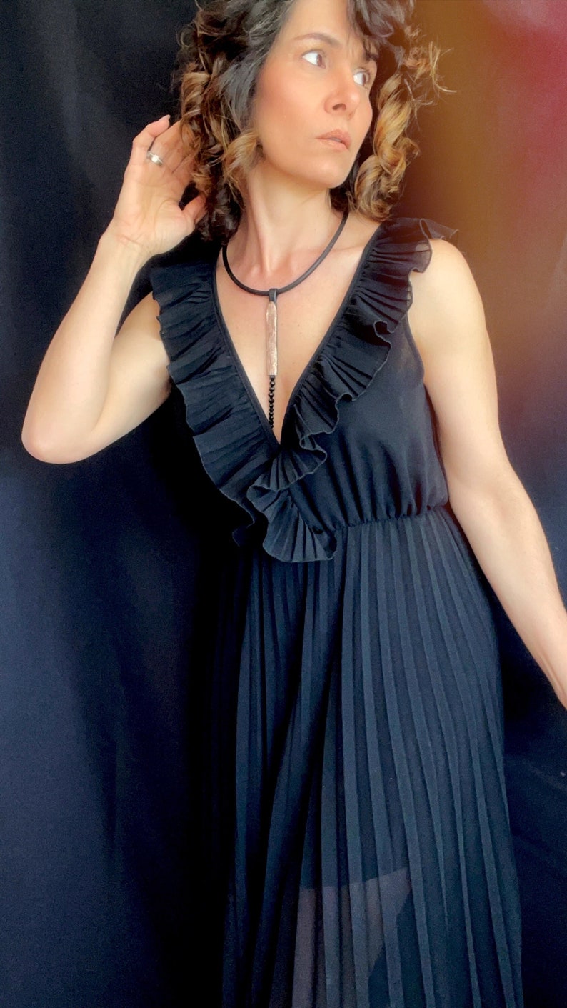 May include: A woman wearing a black sleeveless dress with a ruffled neckline and a long pendant necklace. The dress has a pleated skirt and is made of a soft, flowing fabric.