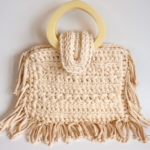 May include: A cream-colored crocheted tote bag with a large, cream-colored, plastic handle and fringe trim.