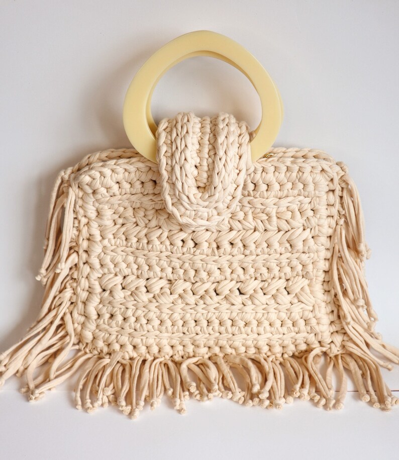 May include: A cream-colored crocheted tote bag with a large, cream-colored, plastic handle and fringe trim.