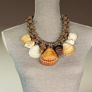 May include: A brown rope necklace with multiple seashells of various sizes and colors. The shells are attached to the rope with knots. The necklace is displayed on a mannequin.