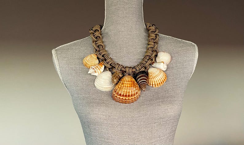 May include: A brown rope necklace with multiple seashells of various sizes and colors. The shells are attached to the rope with knots. The necklace is displayed on a mannequin.