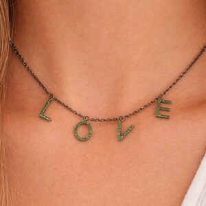 Può includere: Una collana a catena in metallo scuro con quattro ciondoli a forma di lettera che compongono la parola LOVE. Ogni lettera è incastonata con piccole pietre verde brillante, creando un effetto scintillante sulla finitura scura della catena.