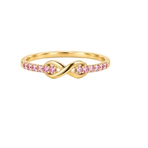 May include: A gold ring featuring a central infinity symbol design. The band is accented with a series of small, round, pink gemstones embedded along both sides of the infinity motif, creating a continuous sparkle across the front of the ring.
