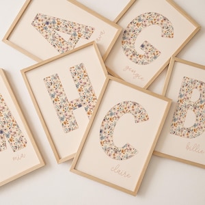 Op de afbeelding: Houten ingelijste bloemenkunstprints. De letters A, B, C, G en H zijn zichtbaar, elk gevuld met een delicaat bloemmotief in tinten roze, blauw en beige. De namen "mia", "ava", "georgie", "claire" en "billie" staan eronder.