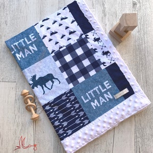 Puede incluir: Una manta para bebé azul y blanca con un diseño de patchwork. La manta presenta un alce, flechas, montañas y las palabras "Little Man".