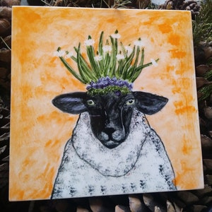 Könnte beinhalten: Quadratische Keramikfliese mit einer skurrilen Illustration eines Schafs mit Blumenkrone. Das Schaf hat ein schwarzes Gesicht und Ohren, weiße Wolle und grüne Augen. Der Hintergrund ist orangefarben strukturiert.