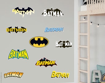 Pode incluir: Um conjunto de nove adesivos de parede do Batman com diferentes versões do logotipo icônico em preto, amarelo e azul. Os adesivos estão dispostos em uma parede cinza claro em um quarto.