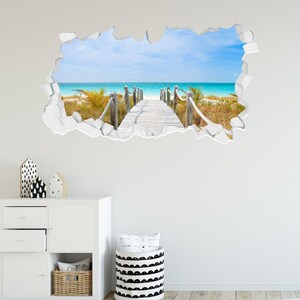 May include: A wall decal featuring a tropical beach scene with a wooden walkway leading to a turquoise ocean. The decal creates the illusion of a hole in the wall, revealing the beach scene.