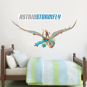 May include: A blue and red dragon with a blonde haired rider flying in the air. The dragon has a large mouth and sharp teeth. The rider is wearing a brown leather jacket and a helmet. The text "AstridStormFly" is printed in blue letters on a white background.