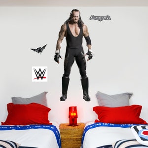 May include: A black and white wall decal featuring a wrestler in a black tank top and black pants. The wrestler is standing with his arms at his sides and has a serious expression on his face. The decal also includes the text "Undertaker" and the WWE logo.