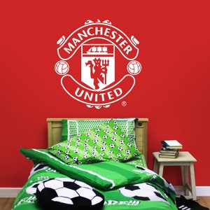 May include: A white Manchester United soccer team logo decal on a red wall above a green and white soccer-themed bed with a white soccer ball pillow.