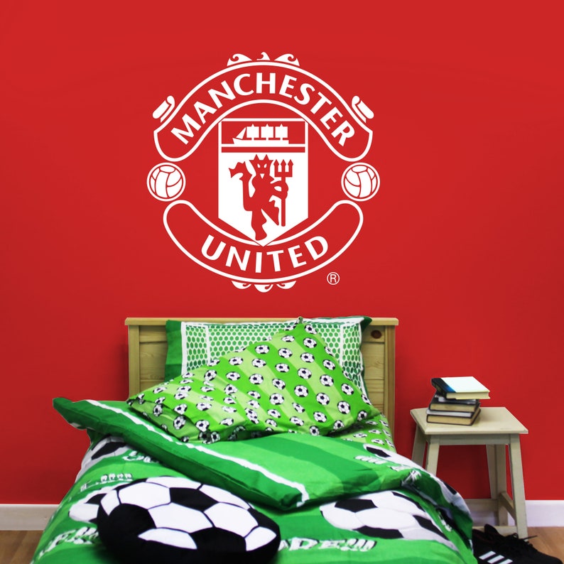 May include: A white Manchester United soccer team logo decal on a red wall above a green and white soccer-themed bed with a white soccer ball pillow.