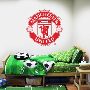 May include: A white wall with a red Manchester United logo decal. A twin-sized bed with green and white bedding featuring soccer balls is in front of the wall. A small wooden table with books and a lamp is next to the bed.