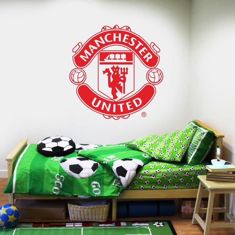 May include: A white wall with a red Manchester United logo decal. A twin-sized bed with green and white bedding featuring soccer balls is in front of the wall. A small wooden table with books and a lamp is next to the bed.