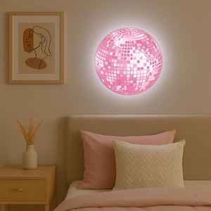 May include: A pink disco ball light fixture on a wall above a bed. The light emits a soft glow. The bed has pink pillows and a beige headboard. A framed art print and a nightstand are also visible in the room.
