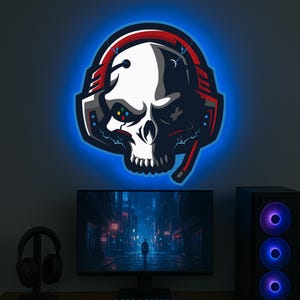 May include: A blue neon light featuring a skull wearing red and black headphones. The skull has a white face with black eyes and a black mouth. The headphones have a red headband and black earcups. The light is on a wall behind a computer monitor and a speaker.