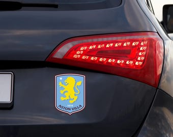以下が含まれることがあります： アストンヴィラの長方形の車用ステッカー。チームのエンブレムが特徴です。エンブレムは、明るい青色の背景に黄色のライオンが描かれており、白い文字で「ASTON VILLA 1874」と書かれています。ステッカーは、暗い灰色の車の後部、テールライトの近くに貼られています。