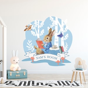 Puede incluir: Una calcomanía de pared azul y blanca con un conejo leyendo un libro en un entorno de bosque. La calcomanía incluye una pancarta con el texto "SAM'S ROOM".