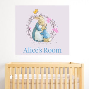 Op de afbeelding: Een lichtpaarse wanddecoratie met een krans van bloemen en twee konijnen die elkaar omhelzen. De tekst "Alice's Room" staat onder de konijnen.