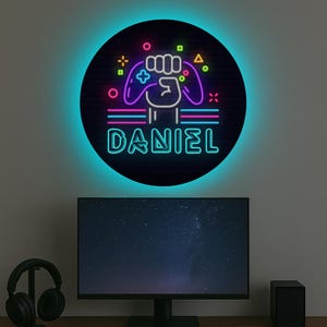 May include: A neon sign featuring a gamer fist holding a controller and the name "DANIEL" in neon lettering. The sign is set against a black background with a blue halo.
