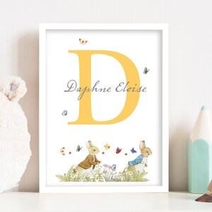 Pode incluir: Uma impressão emoldurada branca com uma letra D amarela e o nome Daphne Eloise escrito em itálico cinza. Dois personagens de Peter Rabbit caminham por um campo de flores com borboletas.