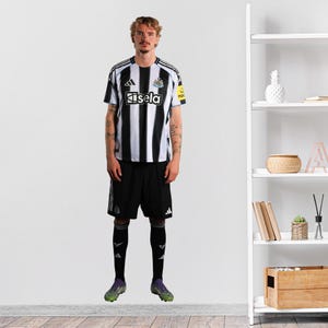 May include: A person in a black and white striped football shirt with the word "Kisela" on the front. They are wearing black shorts, black socks, and green and black football boots. The shirt has a yellow patch on the right sleeve.