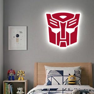Pode incluir: Decoração de parede com o logotipo Transformers vermelho e branco iluminado. O logotipo é montado em uma parede cinza acima de uma estrutura de cama de madeira. Uma imagem emoldurada e robôs de brinquedo também são visíveis.