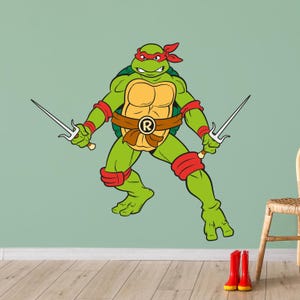 May include: A green Teenage Mutant Ninja Turtle character with a red mask and bandana, holding two sai weapons. The turtle is wearing a brown belt with a large letter 'R' on it.
