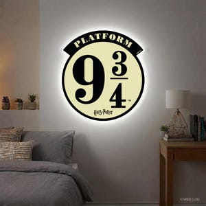 Puede incluir: Una lámpara de pared circular con las palabras "PLATFORM" y "9 3/4" en negro. El texto "Harry Potter" está debajo. La luz tiene un fondo color crema y un borde negro, con un brillo suave. Un artículo decorativo.