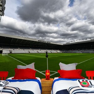 Puede incluir: Dos camas individuales con ropa de cama a rayas blancas y azules y almohadas rojas est&aacute;n instaladas frente a un campo de f&uacute;tbol verde. El campo est&aacute; rodeado por un gran estadio con las palabras "NEWCASTLE UNITED" en el lateral. El cielo est&aacute; nublado.