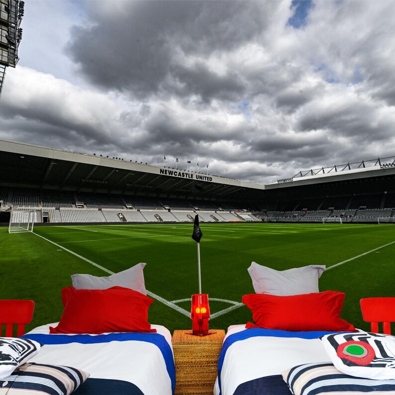 Puede incluir: Dos camas individuales con ropa de cama a rayas blancas y azules y almohadas rojas est&aacute;n instaladas frente a un campo de f&uacute;tbol verde. El campo est&aacute; rodeado por un gran estadio con las palabras "NEWCASTLE UNITED" en el lateral. El cielo est&aacute; nublado.
