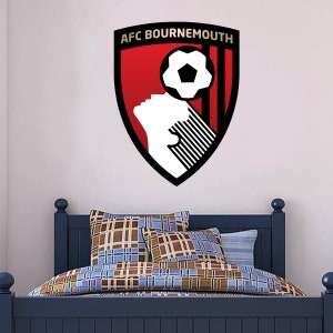 Può includere: Un adesivo murale con lo stemma dell'AFC Bournemouth. L'emblema a forma di scudo presenta uno sfondo rosso, un pallone da calcio bianco e la scritta "AFC Bournemouth". L'adesivo è esposto sopra un letto blu con biancheria da letto fantasia.