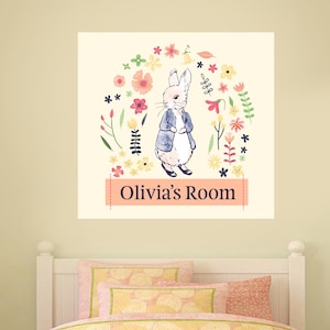 Op de afbeelding: Een wit konijn met een blauw jasje staat in het midden van een cirkel van kleurrijke bloemen. De tekst "Olivia's Room" staat op een roze banner onder het konijn.