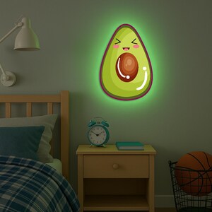 May include: A wall-mounted, glowing avocado-shaped light with a smiling face. The light has a green outer edge, a pink border, and a brown seed. The light is above a wooden bedside table in a bedroom.