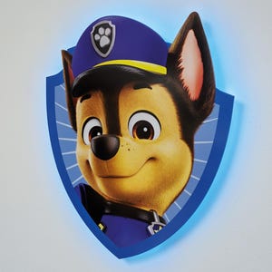Peut inclure: Un personnage de Pat'Patrouille, Chase, est représenté dans un cadre bleu en forme de bouclier. Chase porte un chapeau de policier bleu avec une bande jaune et un badge jaune avec une empreinte de patte. Il a une fourrure brune, un nez noir et une expression amicale.