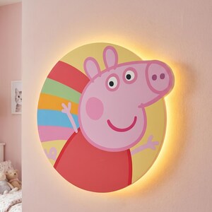 Könnte beinhalten: Runde, beleuchtete Wanddekoration mit einer Cartoon-Figur eines Schweins in einem roten Kleid und einem Regenbogen-Streifenmuster. Das Schwein ist rosa mit einem lächelnden Gesicht und ausgestreckten Armen. Der Hintergrund ist gelb, und das Licht hat einen warmen Schein.