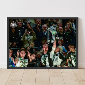 May include: A group of people celebrating with a trophy. The trophy is silver with black and green accents. The people are wearing black and white uniforms with green accents. The trophy has the text "Carabao Cup Final" on it.