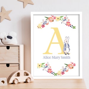 May include: A yellow letter A with a floral wreath around it. The wreath is made of pink, yellow, and blue flowers and green leaves. There are two Peter Rabbit characters in the wreath. The text "Alice Mary Smith" is below the letter A.