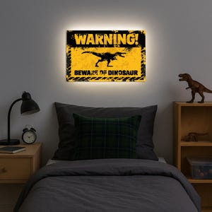 May include: Illuminated wall art featuring a black dinosaur silhouette on a yellow background with the text "WARNING! BEWARE OF DINOSAUR". The sign has a distressed, grunge style and is mounted above a bed.