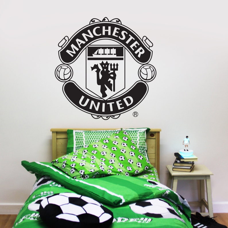 May include: Black and white Manchester United logo on a white wall above a green and white soccer-themed bedspread with a soccer ball pillow.